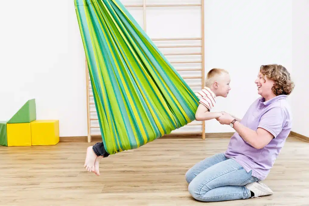 Eine Frau im lila Shirt hält einen Jungen mit den Händen fest, der sich in einer grünen Tuchschaukel befindet.
