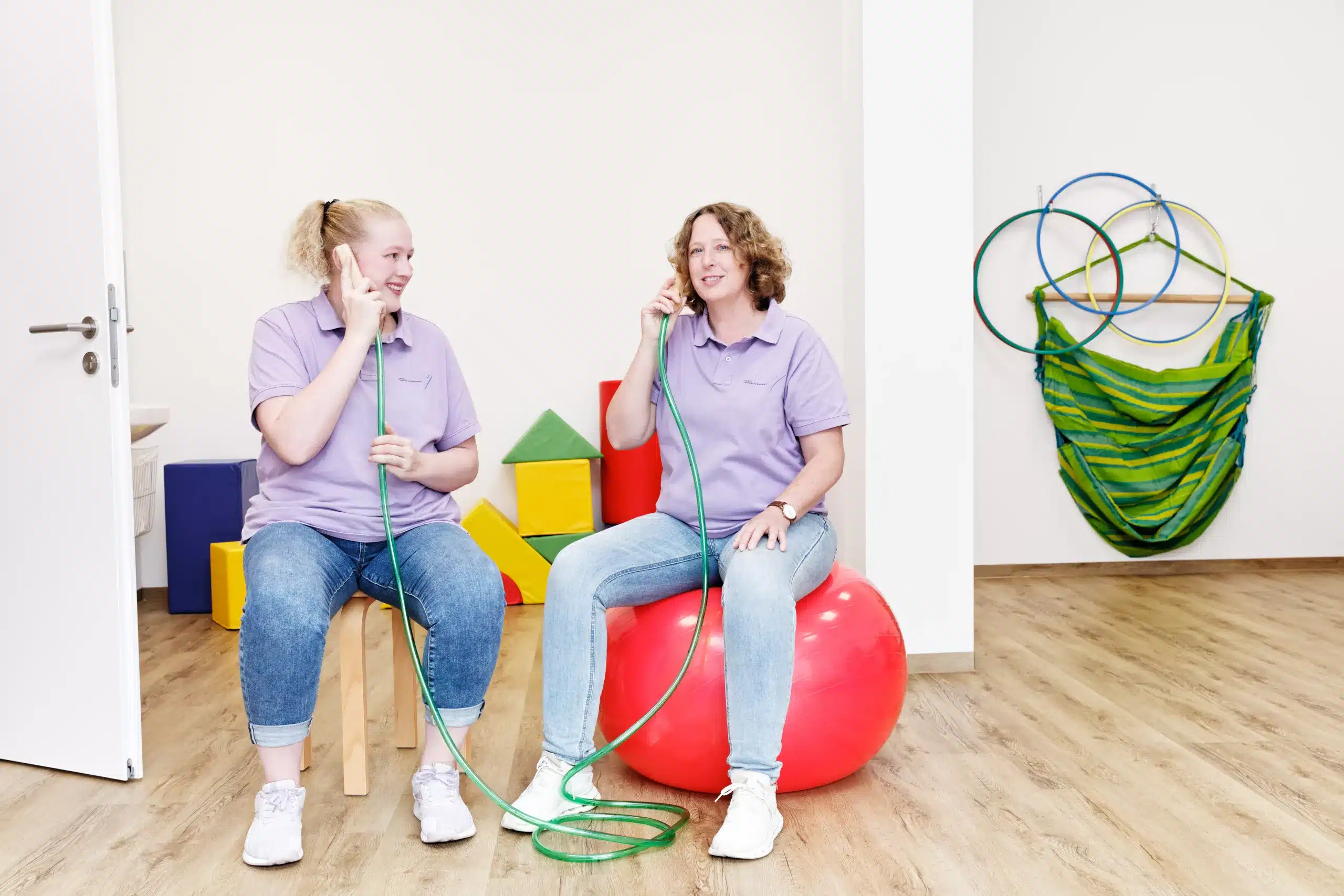 Zwei Frauen im lila T-Shirt sitzen jeweils auf einem Pezziball. Sie halten ein Spielzeugtelefon an die Ohren.